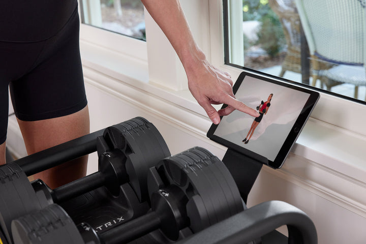 Female selecting workout on tablet beside Matrix dumbbells with side angle close up showing connected strength training setup