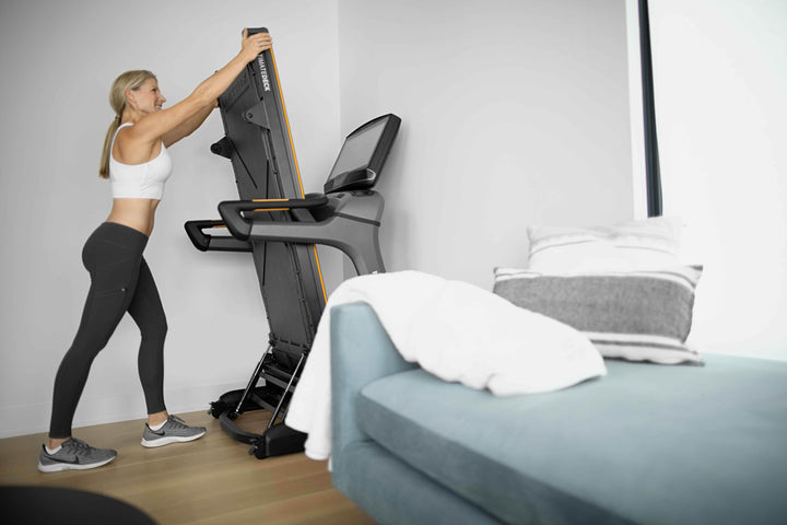 Woman using Matrix folding treadmill in home setting with side angle view showing compact design and cardio workout use