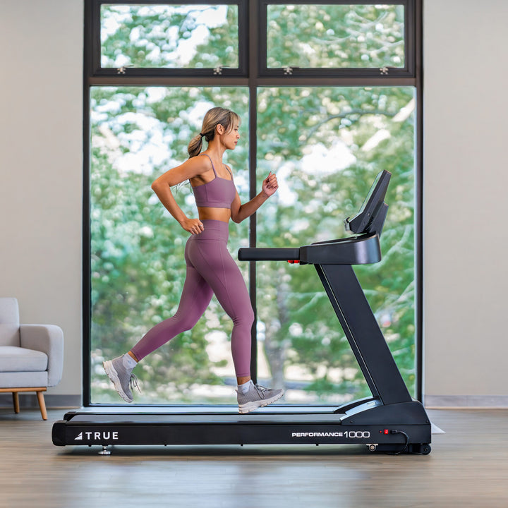 Woman running on a TRUE Performance 1000 treadmill in a bright indoor home workout setting.