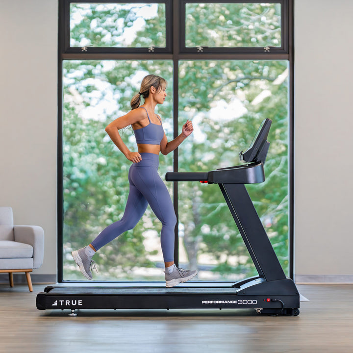 Woman running on a TRUE Performance 3000 treadmill in a bright indoor home workout setting.