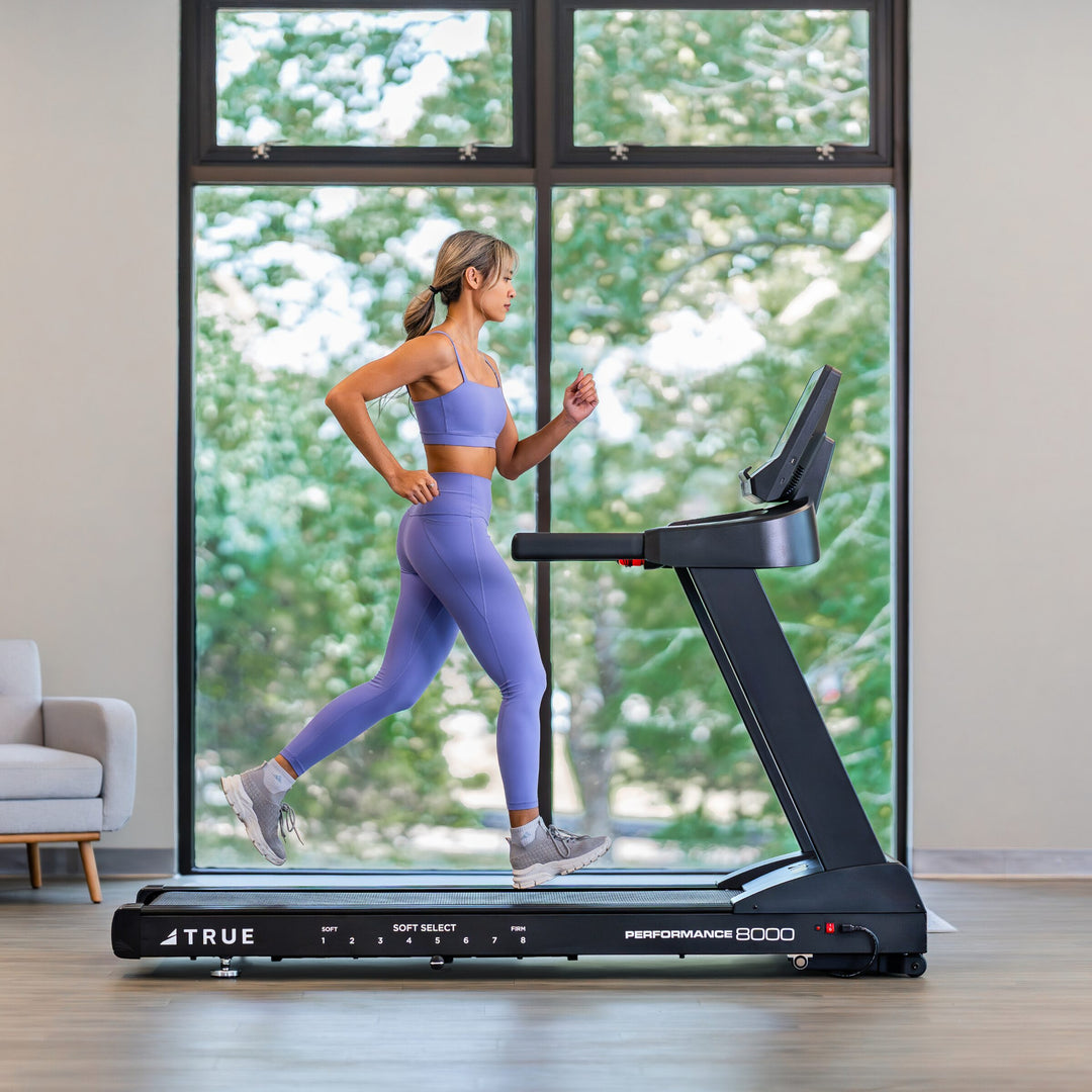Woman running on a TRUE Performance 8000 treadmill in a bright indoor home workout space.