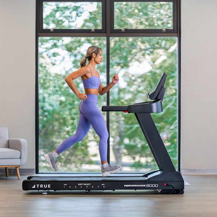 Woman running on a TRUE Performance 8000 treadmill in a bright indoor home workout space.
