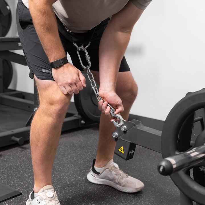 Person connecting a chain to the black Torque USA X-CREATE belt squat attachment before performing a squat exercise.